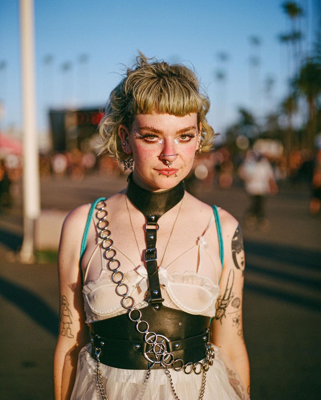 Blonde woman with a black leather harness stands in an outdoor event area with a crowd and palm trees in the background.