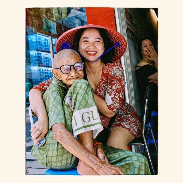 Elderly woman and young woman embrace, both smiling, in front of a store with seating visible in the background, sunny day.