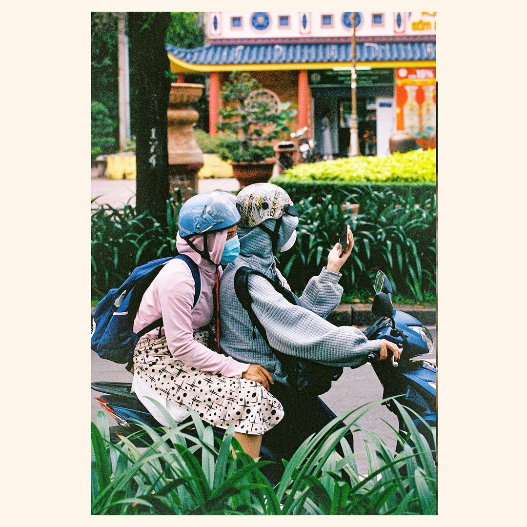 Two people on a scooter in an urban environment. One person is taking a selfie with a smartphone in a film-style photograph.