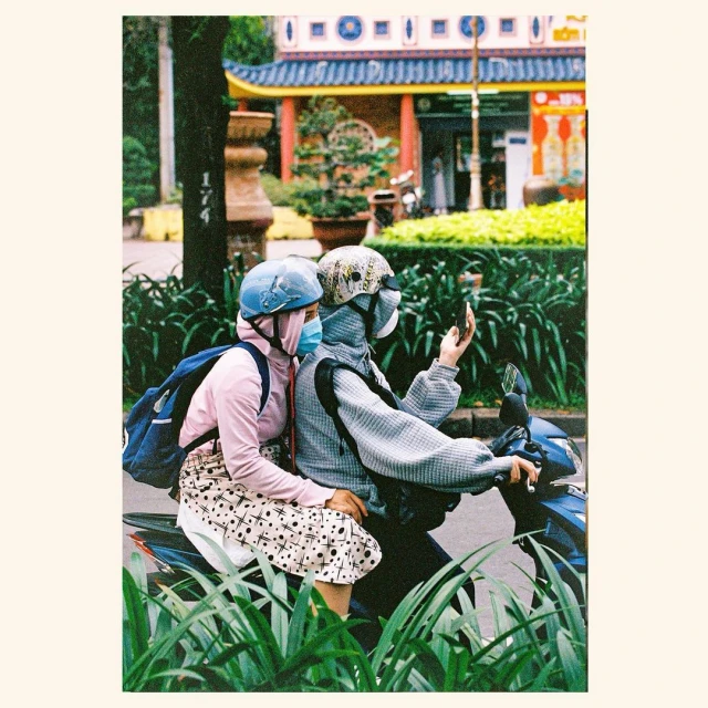 Two people on a scooter in an urban environment. One person is taking a selfie with a smartphone in a film-style photograph.