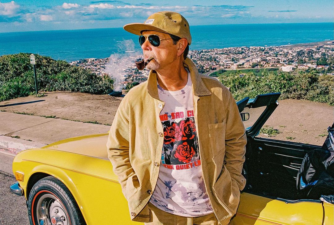 Adult male in corduroy jacket and hat smoking a pipe next to yellow convertible car on hillside overlooking ocean and city.
