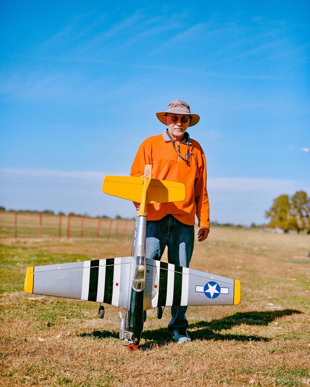 An older adult man poses outdoors on a grassy field, holding a model P-51 Mustang airplane against a clear blue sky backdrop.