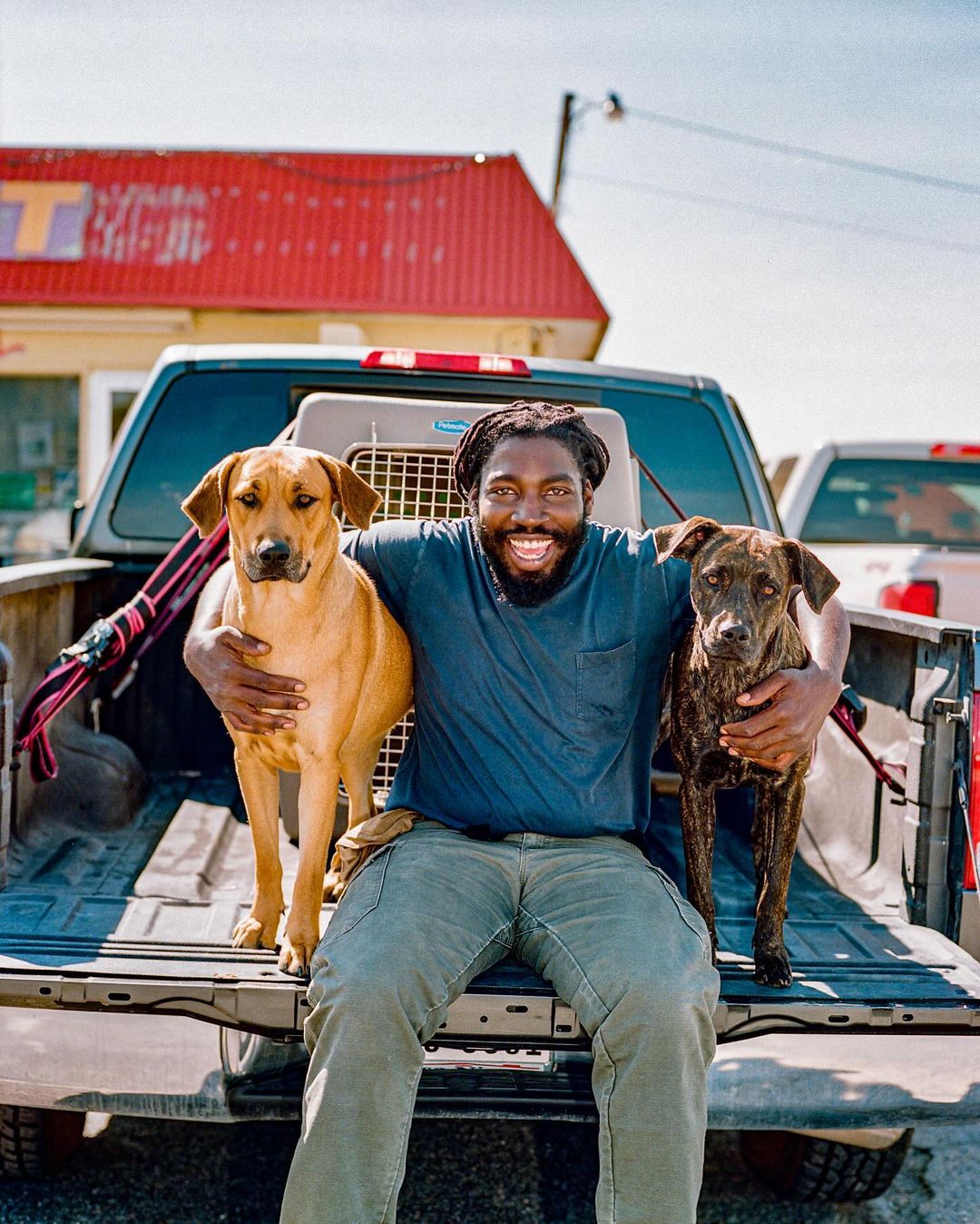 A man sits in a pickup truck with two dogs; one tan and the other a darker brindle. All are smiling on a bright day outdoors.