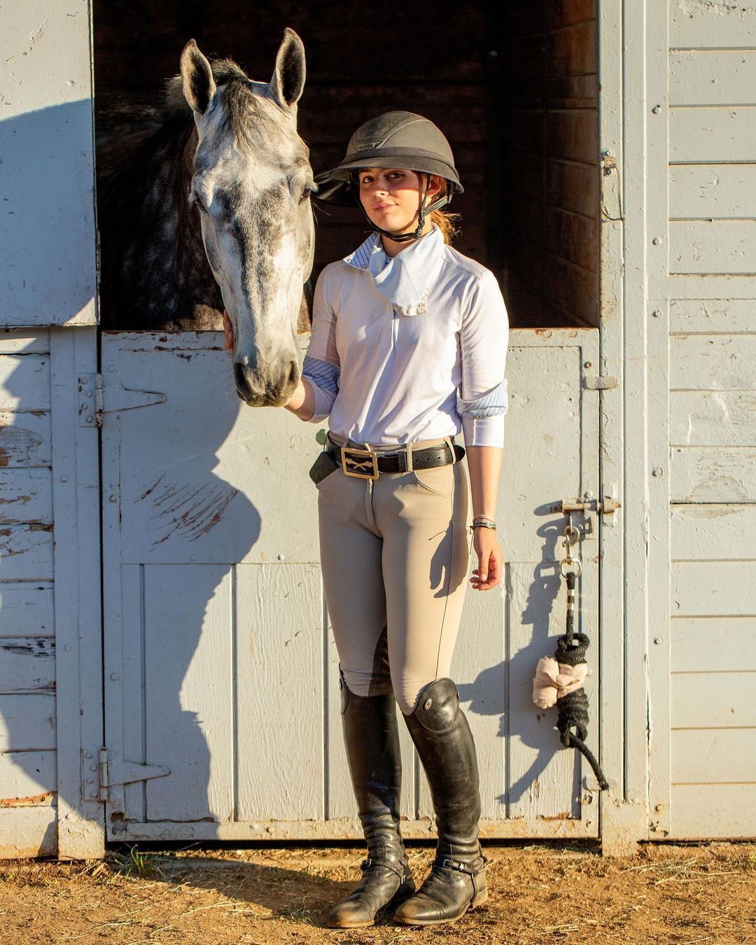 Young woman in riding attire poses with her dappled grey horse in a stable doorway, bathed in soft sunlight. Equestrian lifestyle.