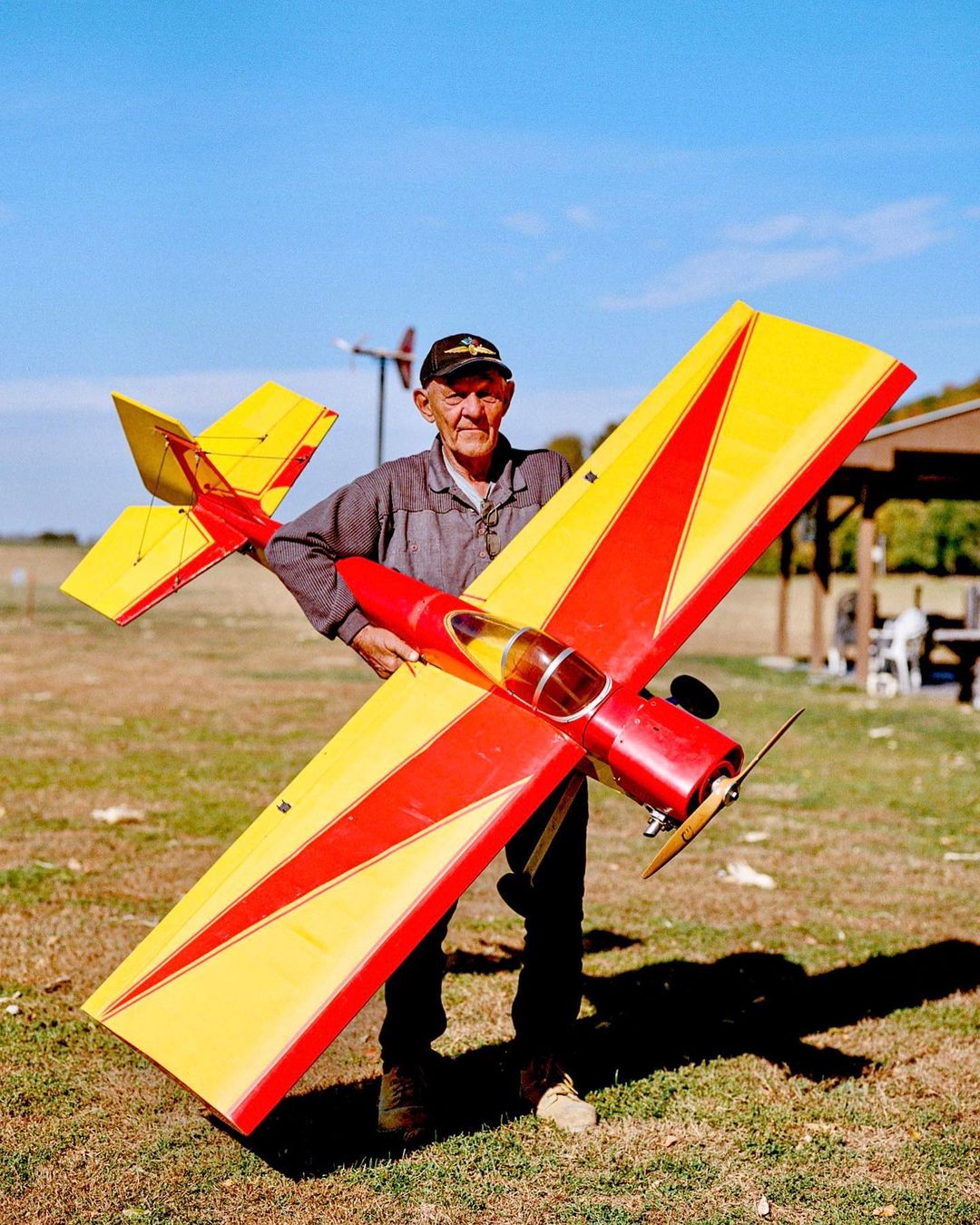 Elderly man smiles, holding yellow and red model airplane outdoors on grassy field under a blue sky, captured in a medium shot.