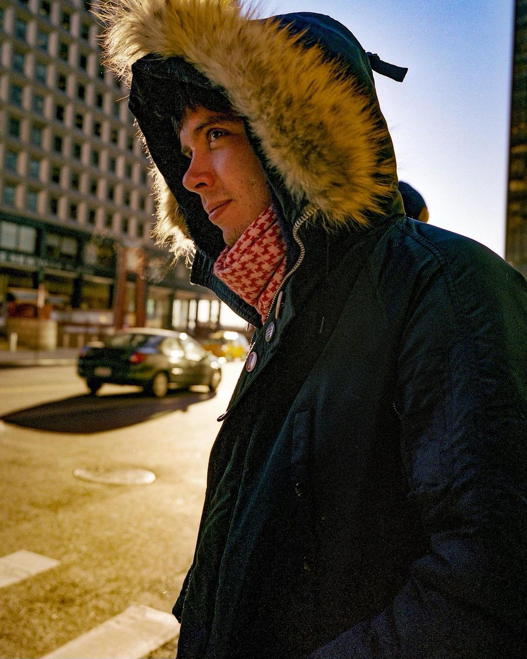 A young adult male wearing a hooded winter coat stands on a city street with buildings in the background on a sunny day.