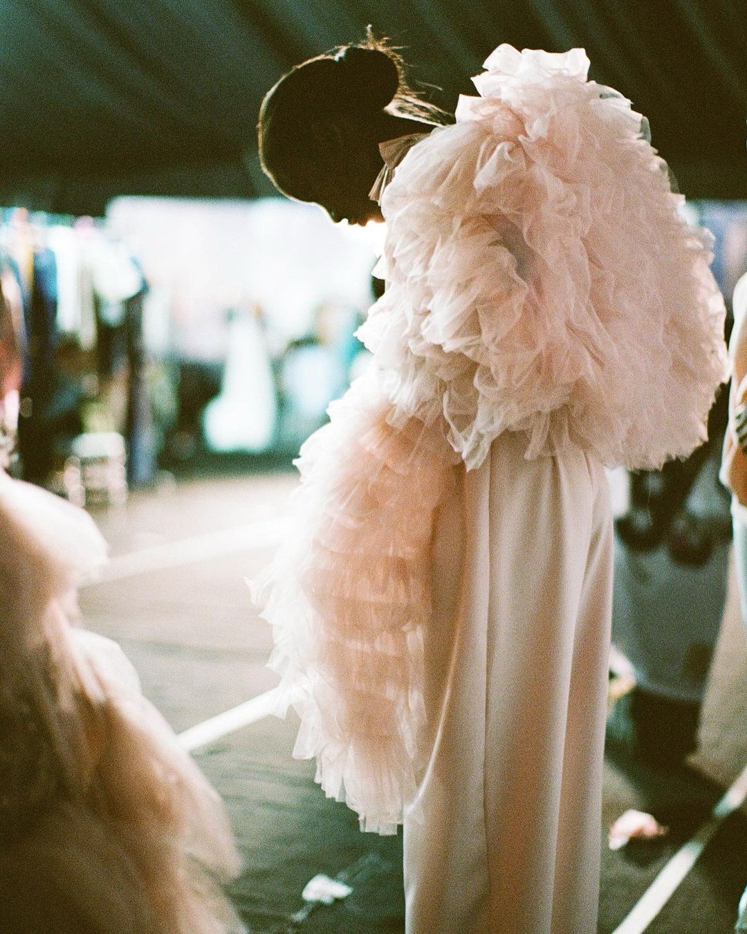 A fashion photo features a person in an elaborate, layered pink ruffle jacket with white pants at what appears to be a backstage venue.