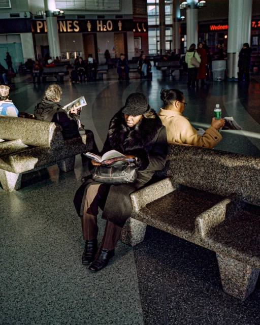 Woman in a coat reading a book while sitting on a stone bench in a train station, with other people and signage in the background.