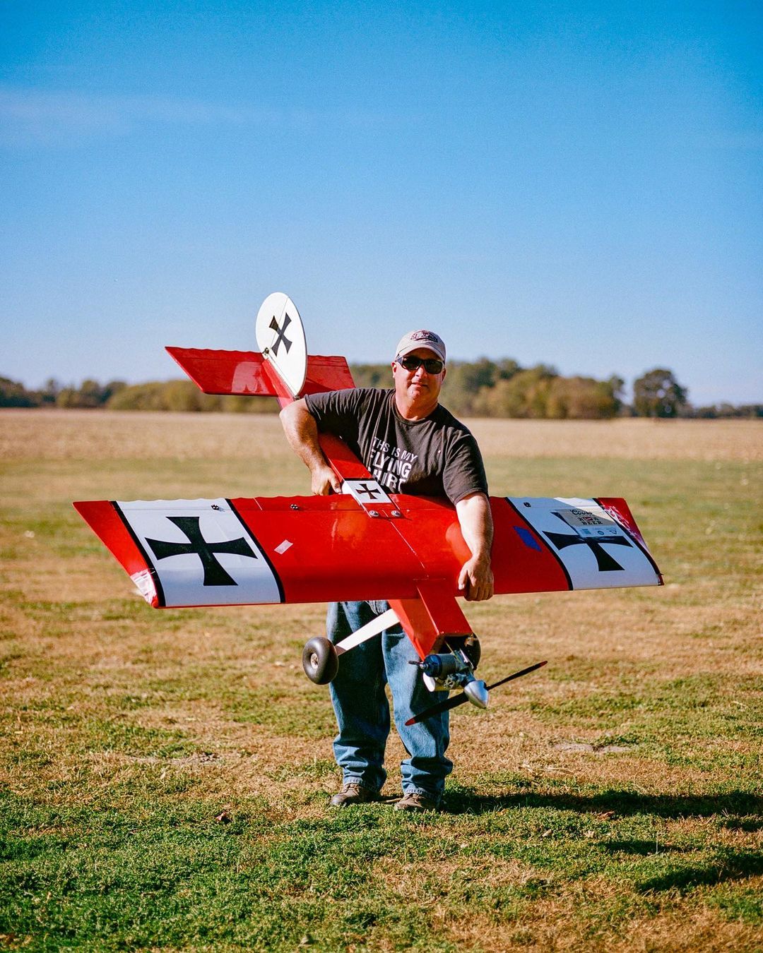 Adult male stands outdoors on grass holding a large red and white model airplane with black crosses, under a clear blue sky.