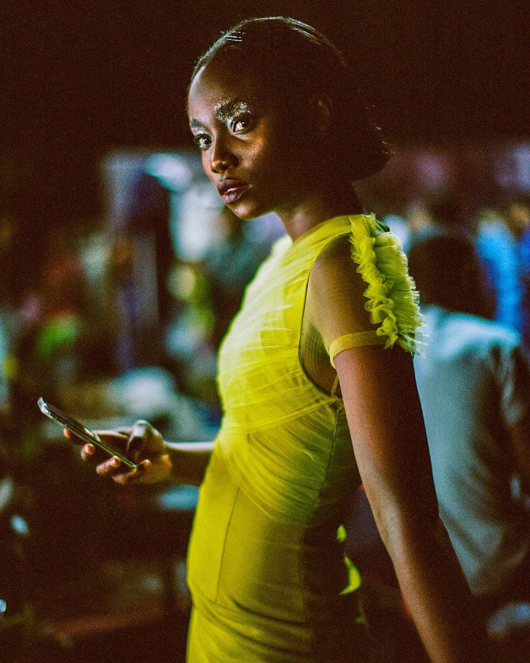 Stylish woman in neon yellow dress looking at the camera while holding a cell phone with people in the background.