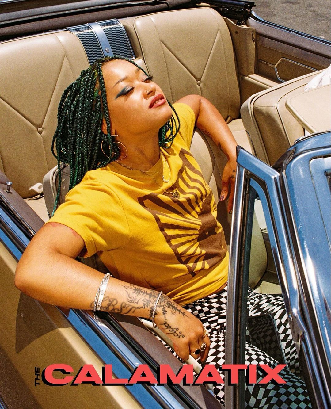 Woman with braided hair poses in a vintage convertible car with a golden paint job, wearing a yellow shirt and patterned pants.