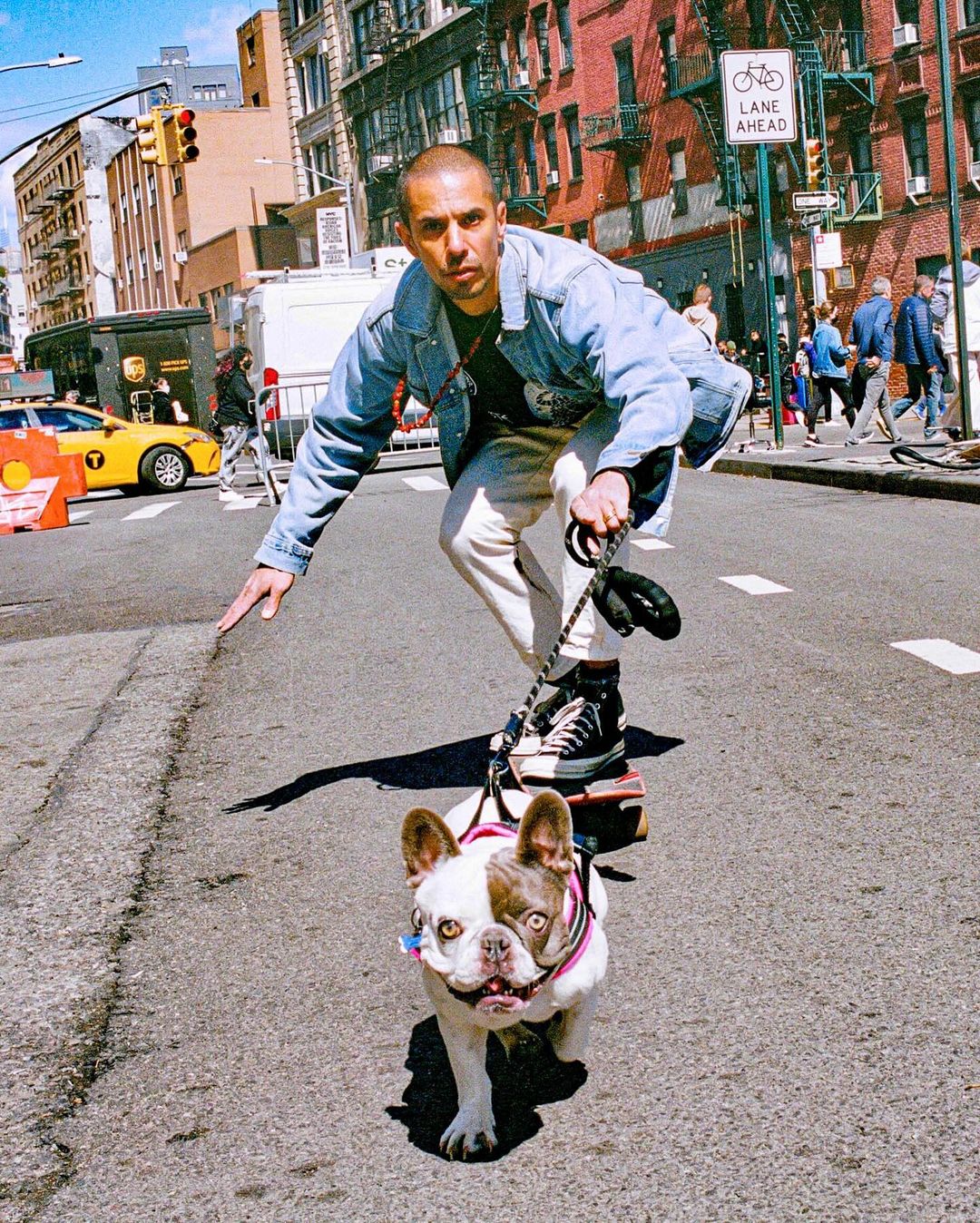 Man rides a skateboard with a dog on a leash down a city street with buildings and a taxi in the background on a sunny day.