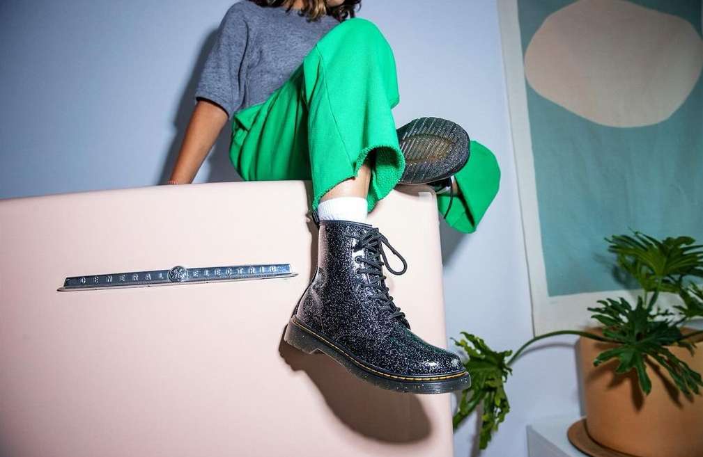 Person in green pants sits on pink refrigerator showcasing black glitter combat boot and modern art print in minimalist interior.