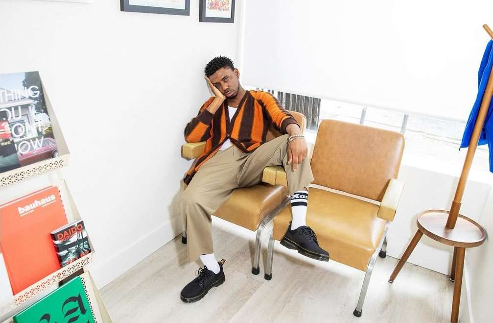 Man sitting in tan chair wearing a striped cardigan and tan pants with books visible in the background, natural light.
