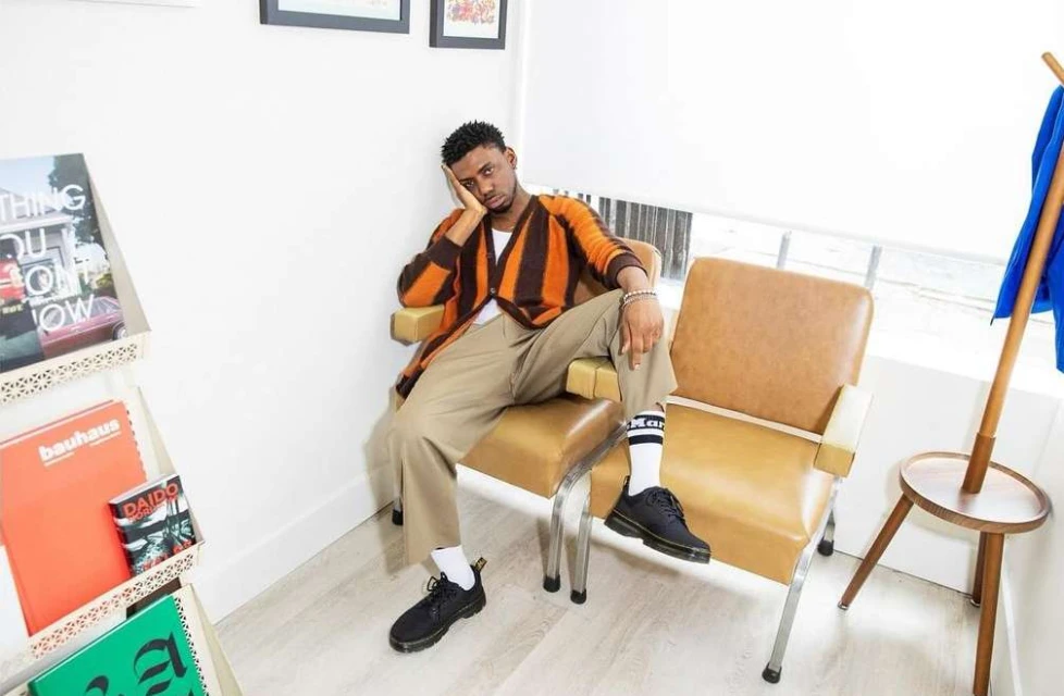 Man sitting in tan chair wearing a striped cardigan and tan pants with books visible in the background, natural light.