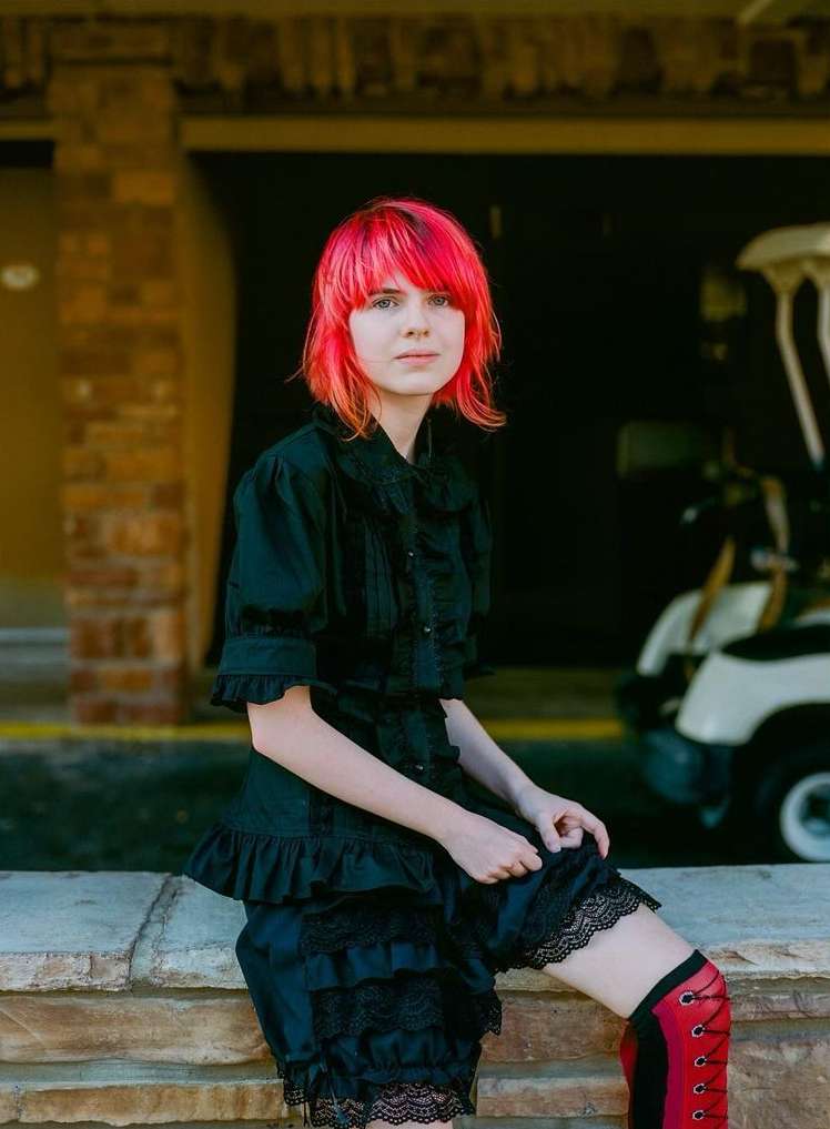A fair-skinned woman with bright pink hair in a black Victorian-style outfit sits on a stone ledge in front of a building with golf carts.