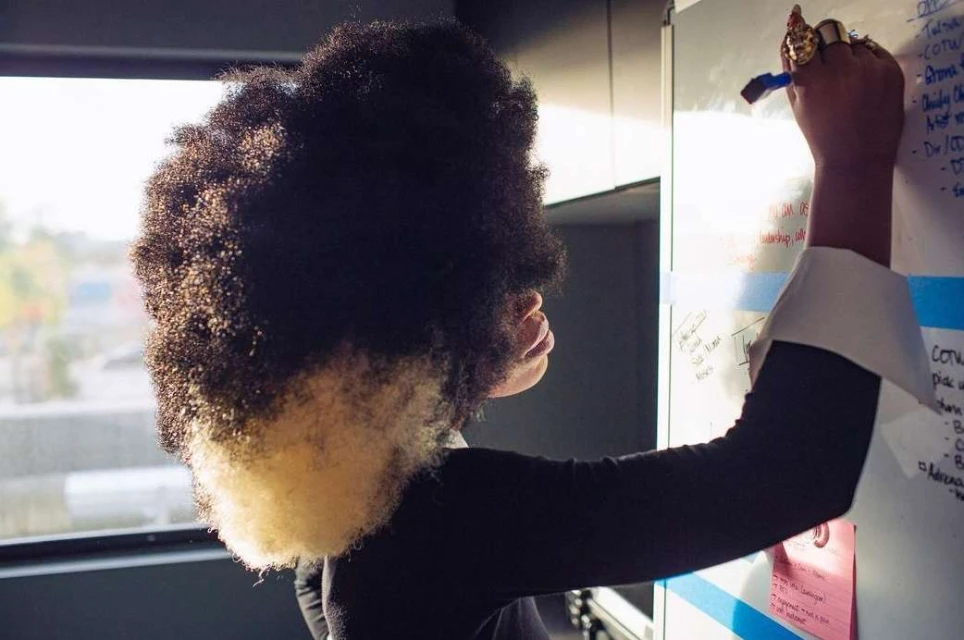 Woman with large afro writing on whiteboard with notes in office setting; blue marker, dark sleeves, and a window are visible.