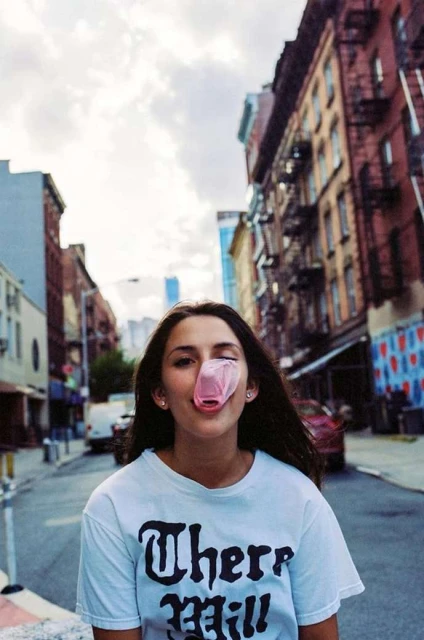Young woman blowing bubble gum bubble on city street with brick buildings and overcast sky in the background.