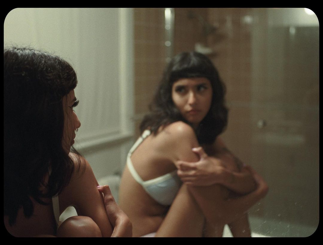 Woman in bra sits near mirror in bathroom, reflecting her pose in muted light with tile and white trim interior.