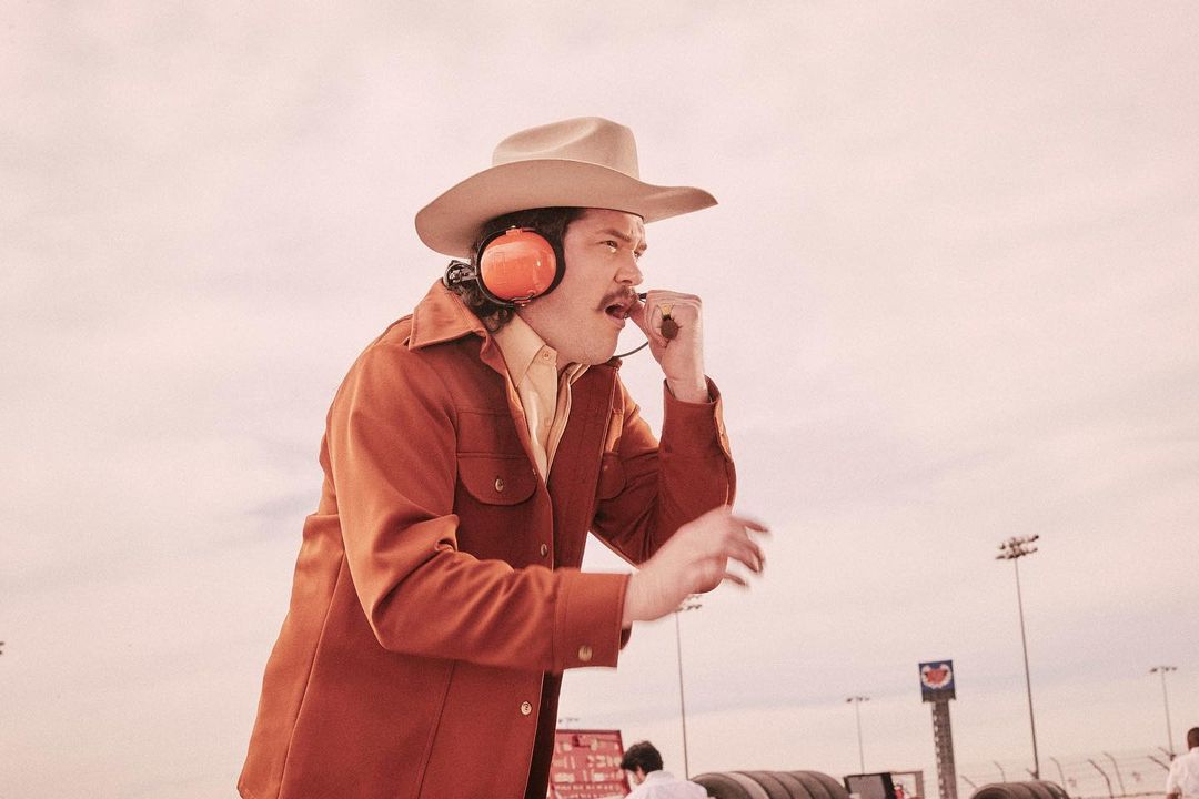 Adult male wearing a cowboy hat and headset speaks with a raised fist at an outdoor car racing event on a sunny day with a clear sky.