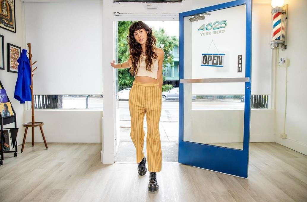 A young adult woman stands in a doorway, exiting a barber shop with an open sign, wearing striped pants and a cream-colored crop top.