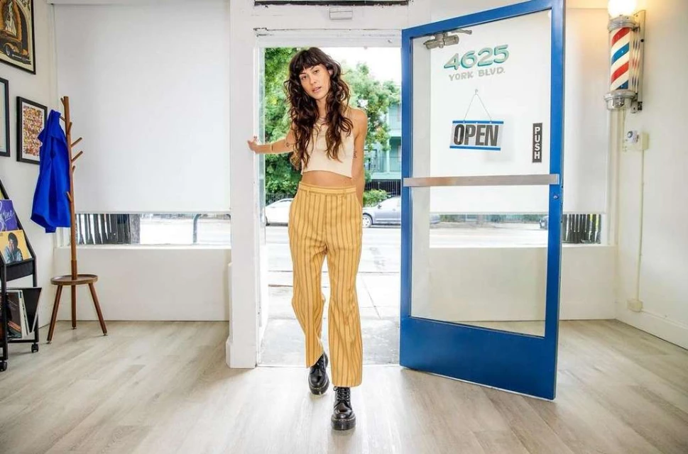 A young adult woman stands in a doorway, exiting a barber shop with an open sign, wearing striped pants and a cream-colored crop top.