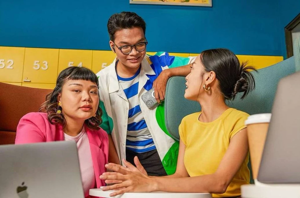 Three young adults collaborate in a modern workspace, two working on laptops and one standing, in a brightly lit, colorful setting.