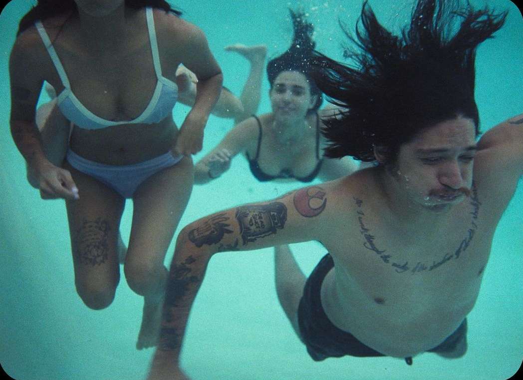 Three people swimming underwater in a bright pool, including a tattooed man in the foreground and two women swimming behind him.
