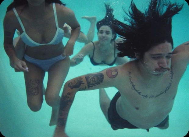 Three people swimming underwater in a bright pool, including a tattooed man in the foreground and two women swimming behind him.