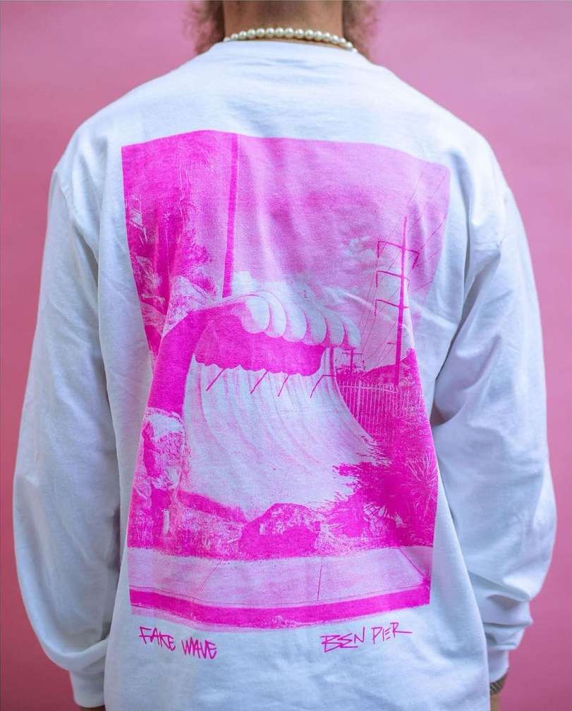 Person wearing a white long-sleeve shirt with a pink graphic of a pier and wave, standing in front of a pink backdrop, pearls.