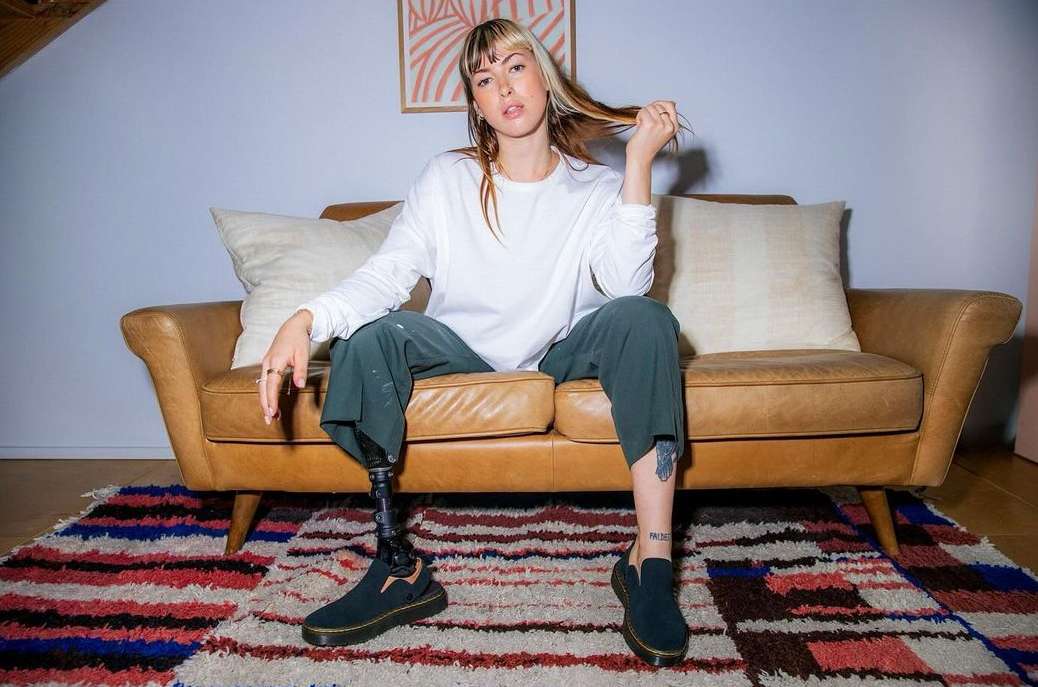 Young woman with prosthetic leg in casual style sitting on a brown leather couch in a living room with a colorful rug.