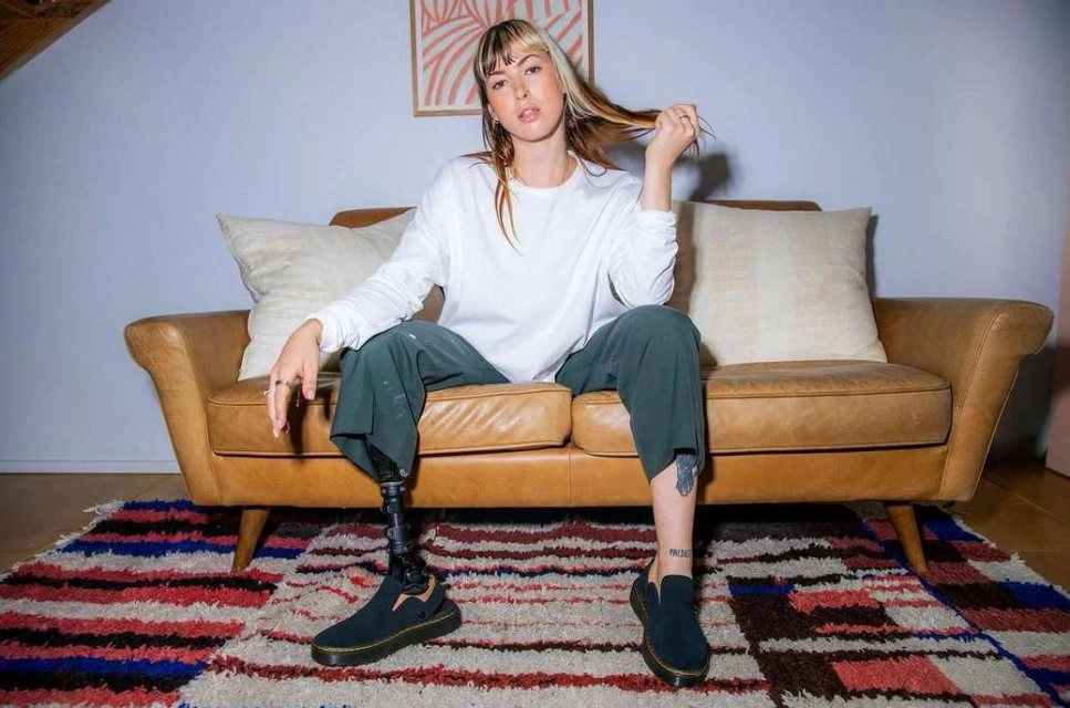 Young woman with prosthetic leg in casual style sitting on a brown leather couch in a living room with a colorful rug.