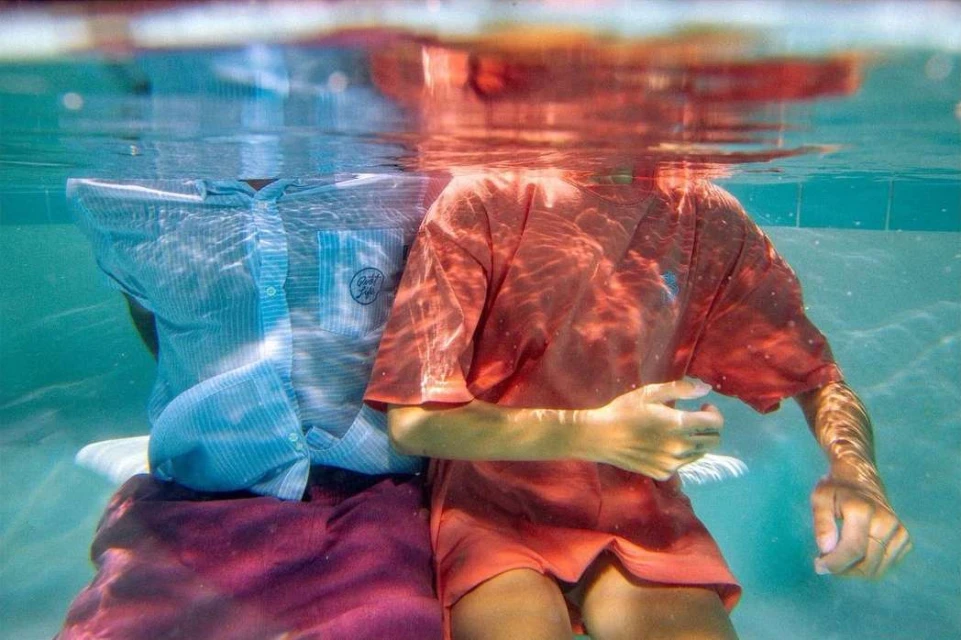 Two people in shirts and skirts underwater in a swimming pool with dappled light and water reflections on their clothes and bodies.