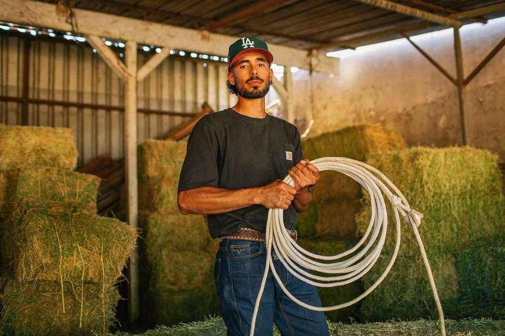 Man in baseball cap holding a coiled white rope in a barn with hay bales in the background, looking at the camera in medium shot.