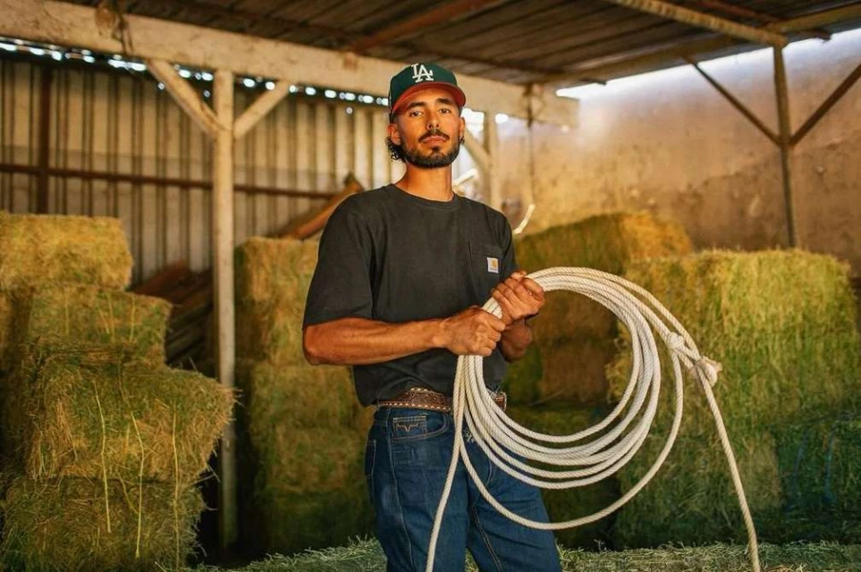 Man in baseball cap holding a coiled white rope in a barn with hay bales in the background, looking at the camera in medium shot.