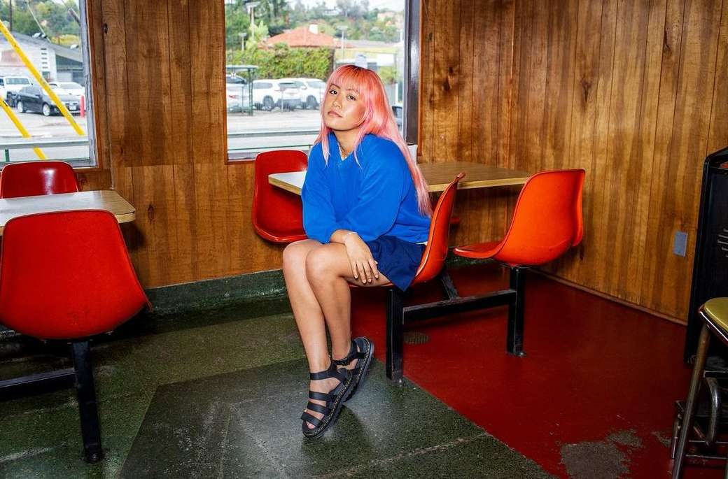 Young woman with dyed pink hair poses while sitting in a vintage diner, wearing a blue sweatshirt and black sandals indoors.