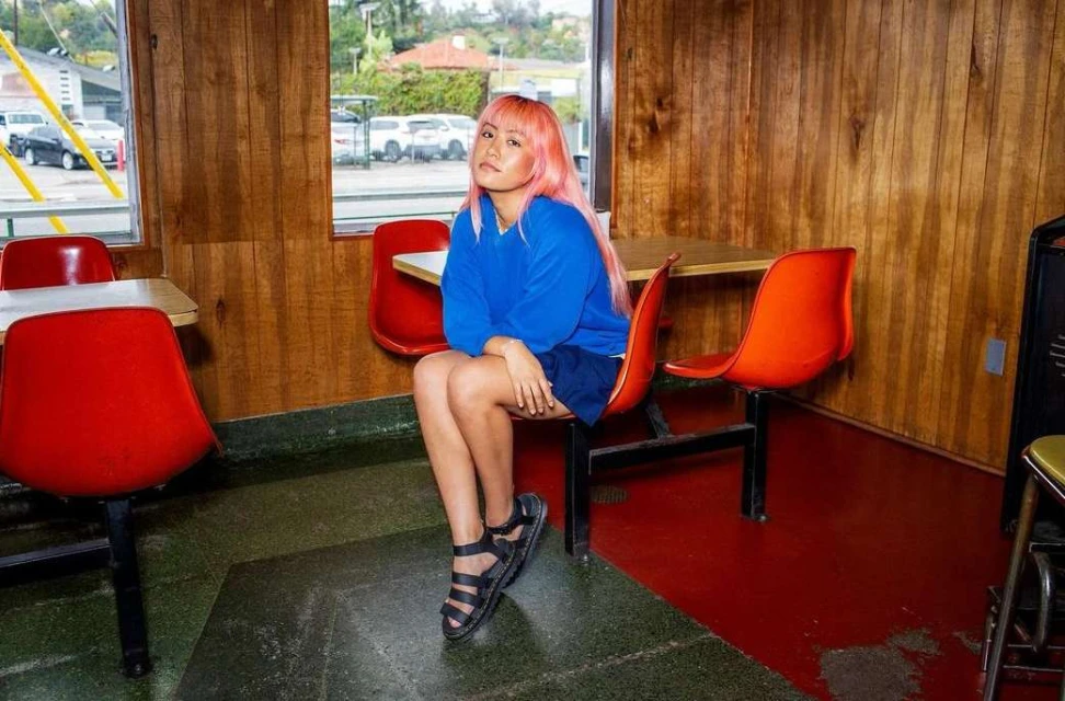 Young woman with dyed pink hair poses while sitting in a vintage diner, wearing a blue sweatshirt and black sandals indoors.