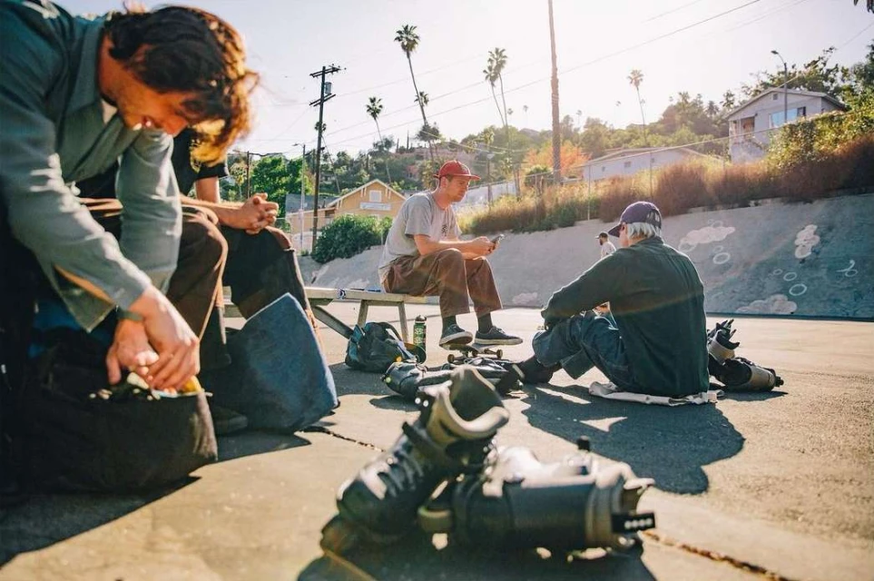 Group of skaters relax in an urban skate park on a sunny day; four adult males, skateboards, and roller blades are visible.