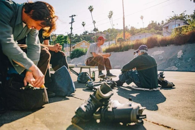 Group of skaters relax in an urban skate park on a sunny day; four adult males, skateboards, and roller blades are visible.