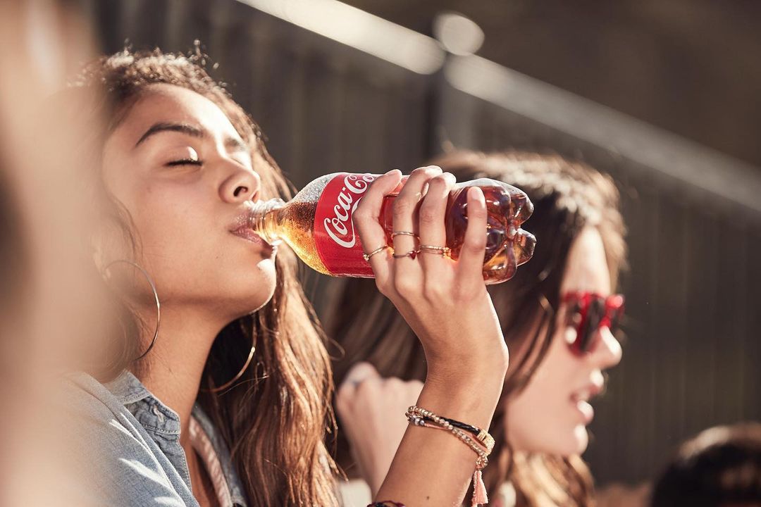 A young woman drinks a Coca-Cola from the bottle. Another female with sunglasses is visible in the background with blurred faces visible.