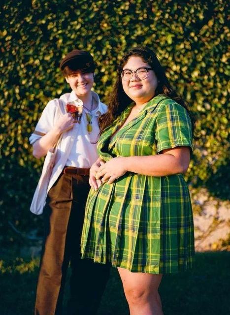 Two people, one in a green plaid dress and the other in a white shirt and brown pants, pose in front of a green leafy wall.