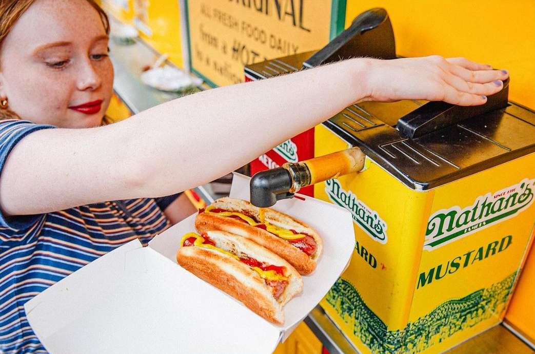 Woman dispenses mustard onto two hot dogs from a Nathan's Famous mustard dispenser. The hot dogs are in a white cardboard tray.
