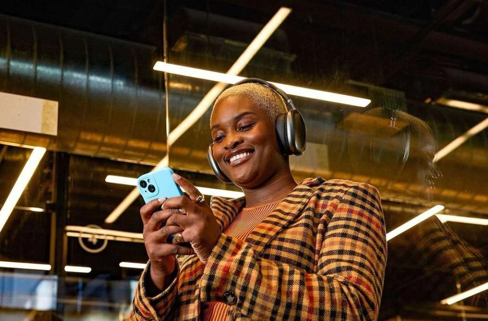 A smiling Black woman with short blonde hair wears headphones and a plaid jacket while holding a blue smartphone in an indoor setting.