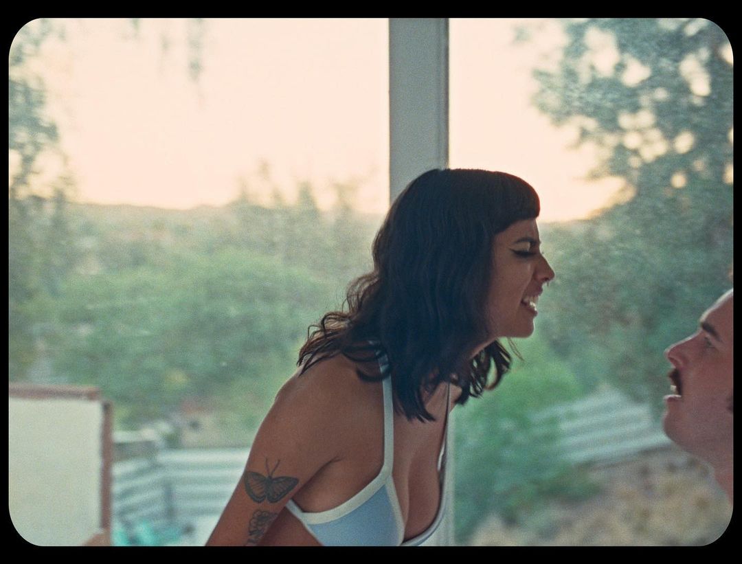Close-up of an adult woman talking to an adult man inside a home with a large window overlooking a green landscape and bright skies.