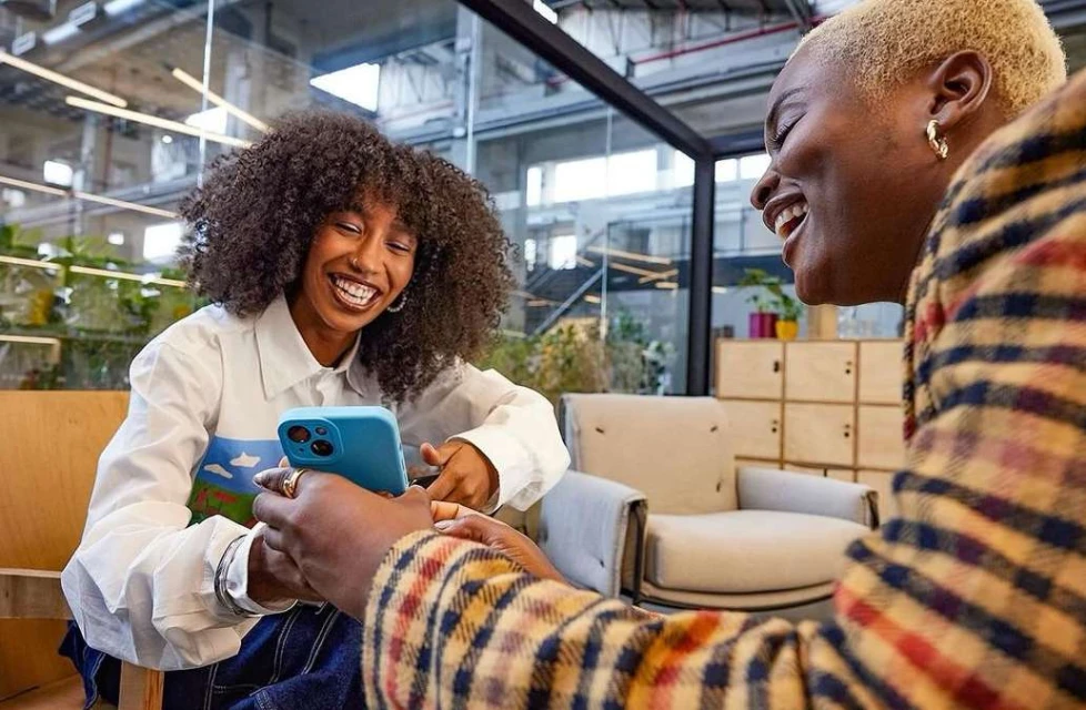 Two happy African-American women laugh while looking at a blue smartphone in an indoor office setting with modern decor and natural light.