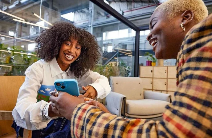 Two happy African-American women laugh while looking at a blue smartphone in an indoor office setting with modern decor and natural light.