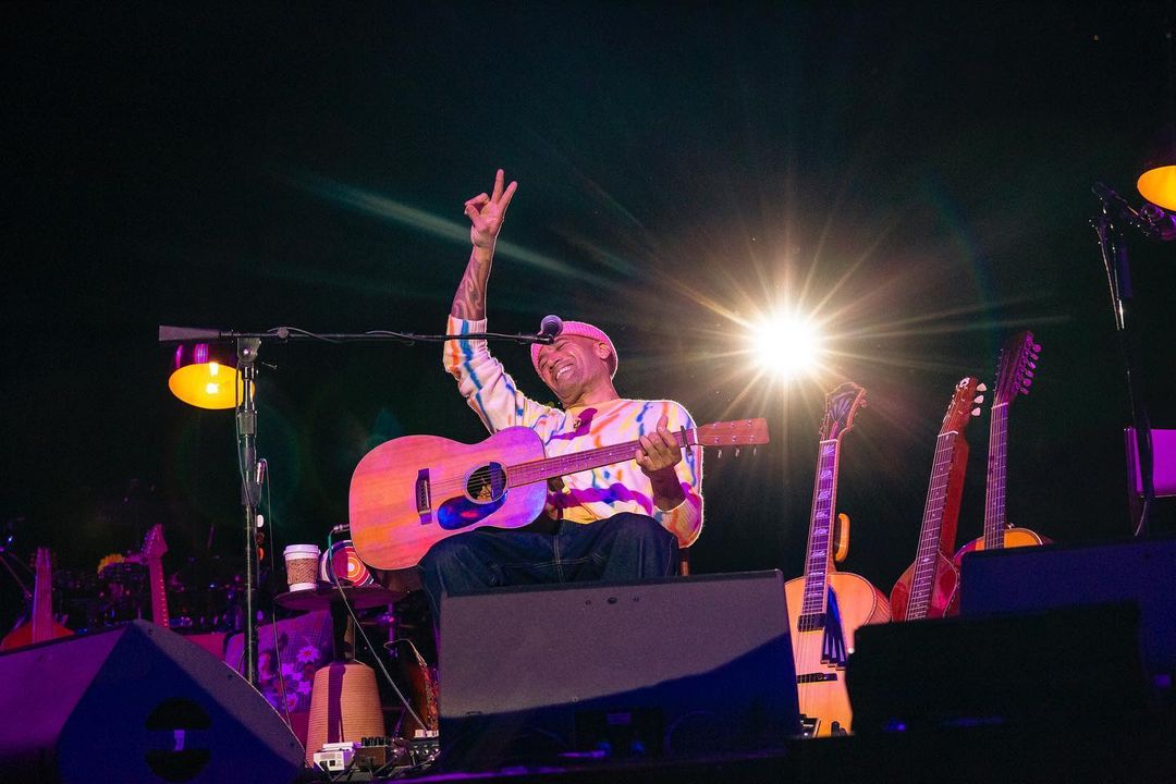 Musician performs on stage playing an acoustic guitar, wearing a colorful shirt, with a peace sign, illuminated by bright stage lights.