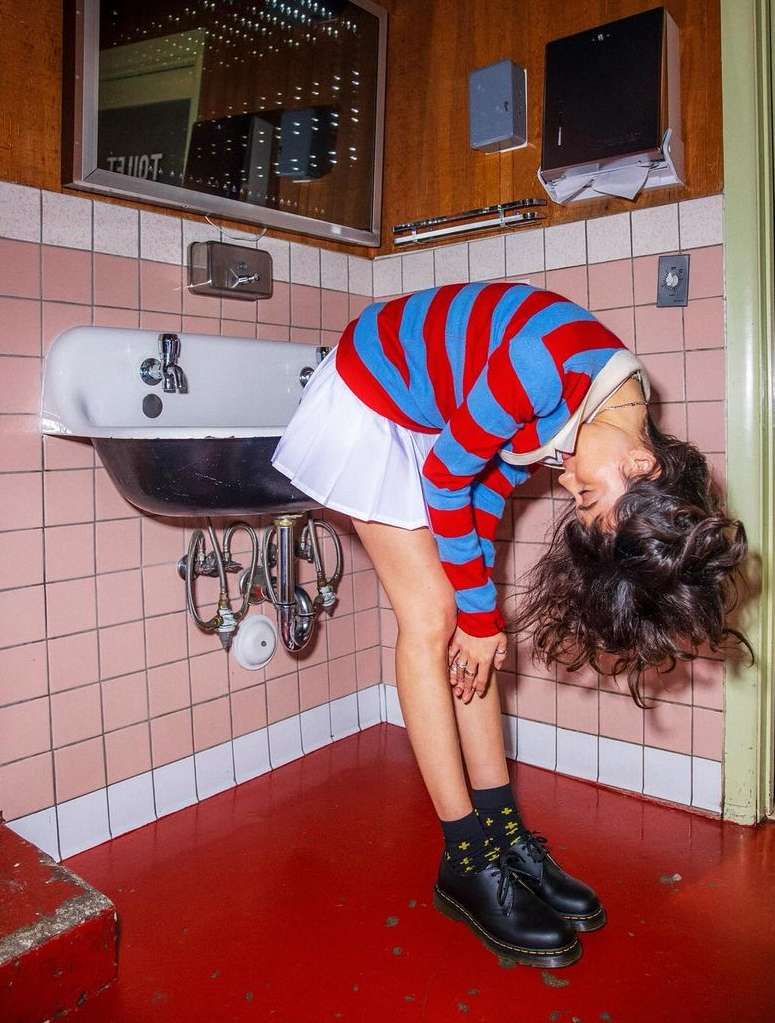 Woman in red and blue sweater bends backward toward a bathroom sink in a pink-tiled retro-style bathroom with a red floor.