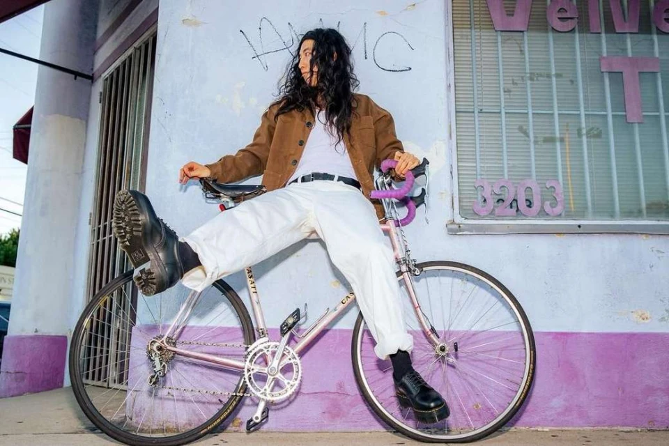 Adult female with long dark hair poses on a pink bicycle in front of an urban building with a purple facade and graffiti.