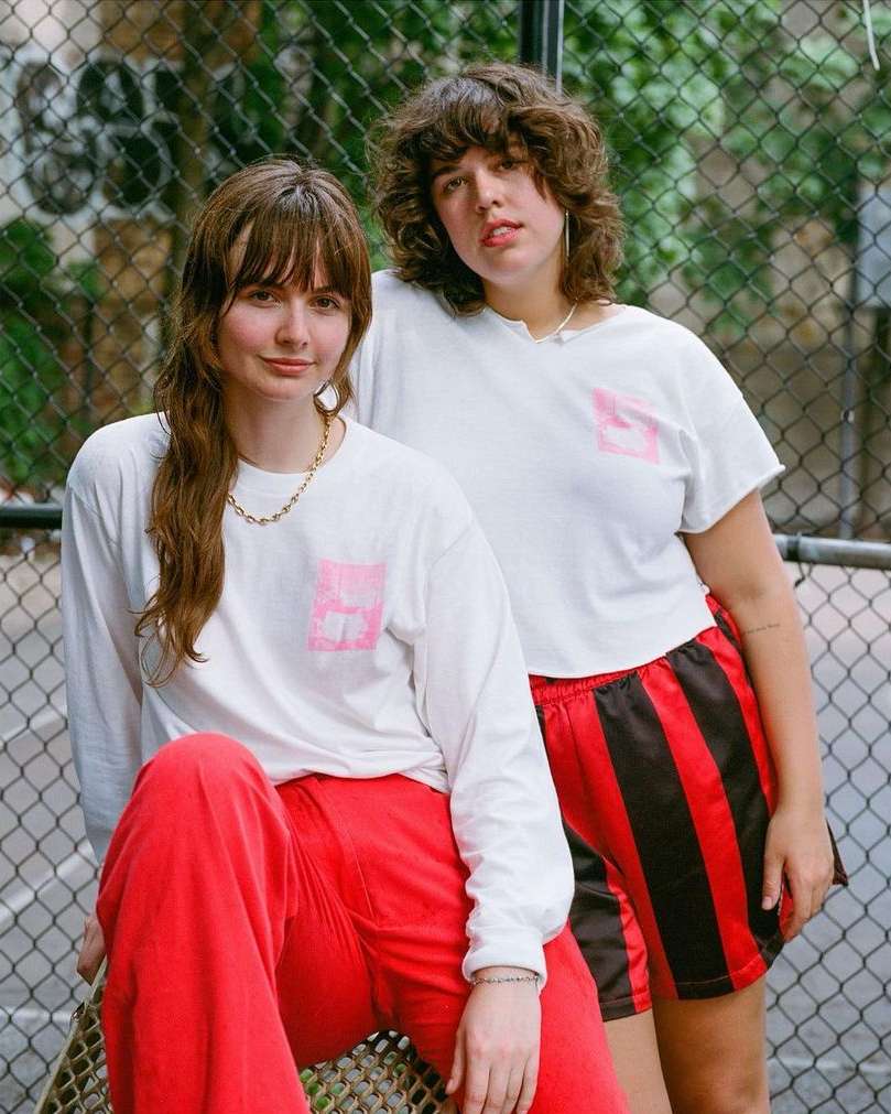 Two young women pose in front of a chain-link fence. One is seated in red pants, the other stands wearing shorts with black stripes.